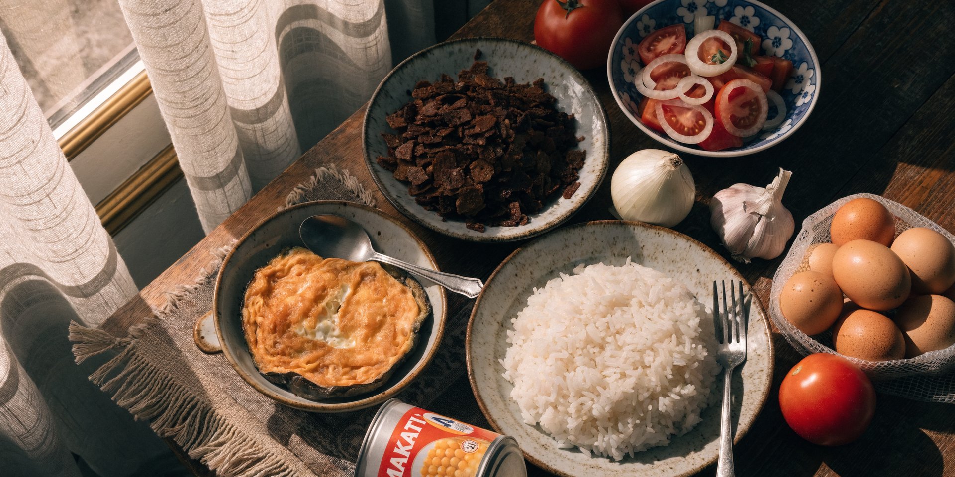 A top-down view of a wooden table featuring Tortang Talong, white rice, adobo flakes, tomato salad, eggs, and canned goods.