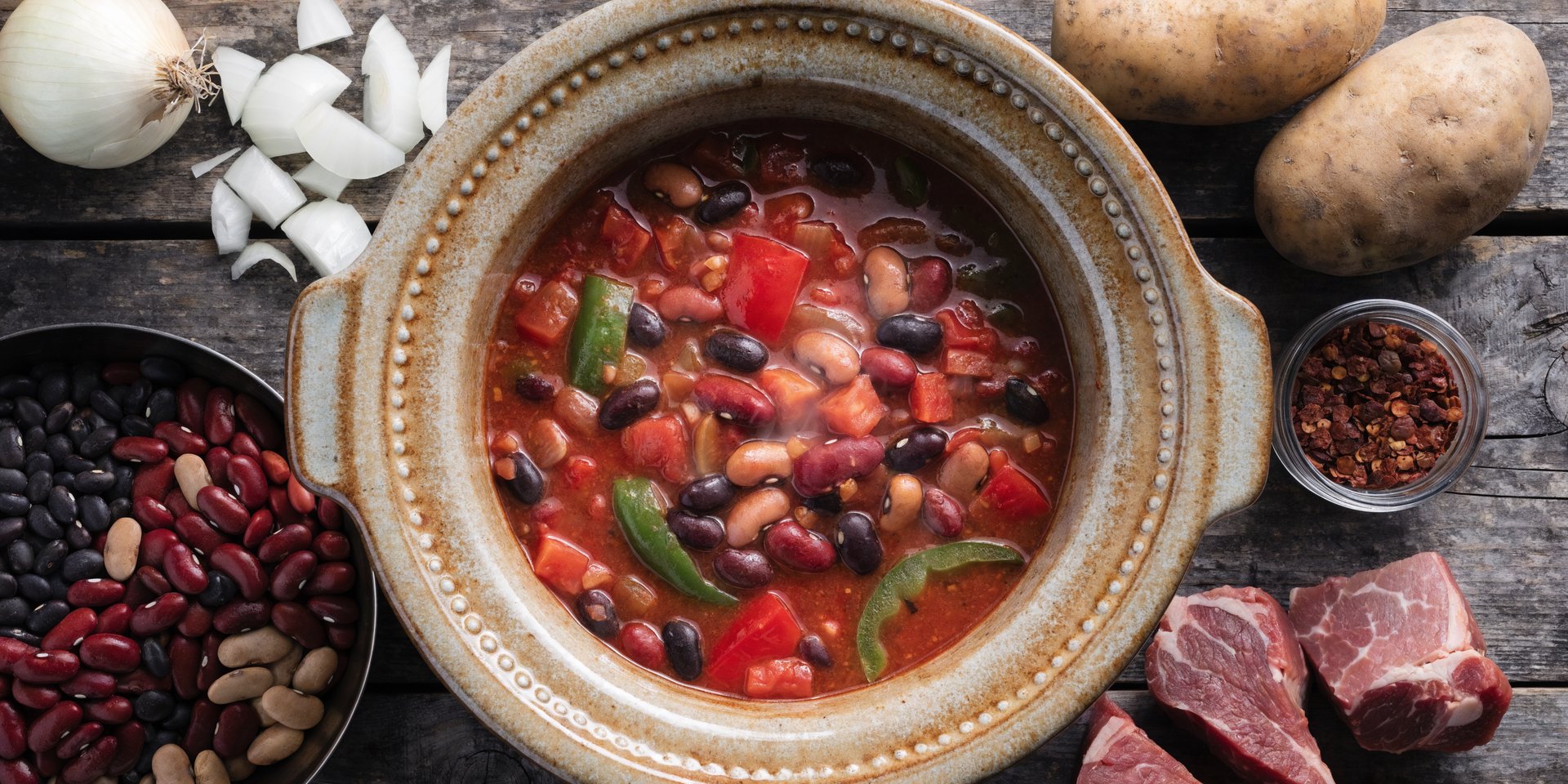 A large slow cooker is filled with a hearty stew, surrounded by dried beans, potatoes, and onions.