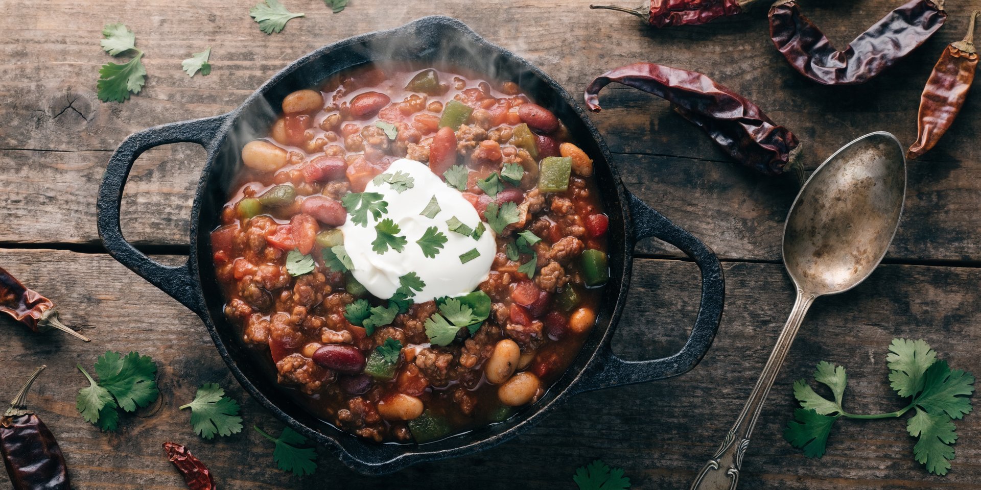 An overhead view of a large, rustic skillet filled with ground beef and bean chili, garnished with cilantro and sour cream.