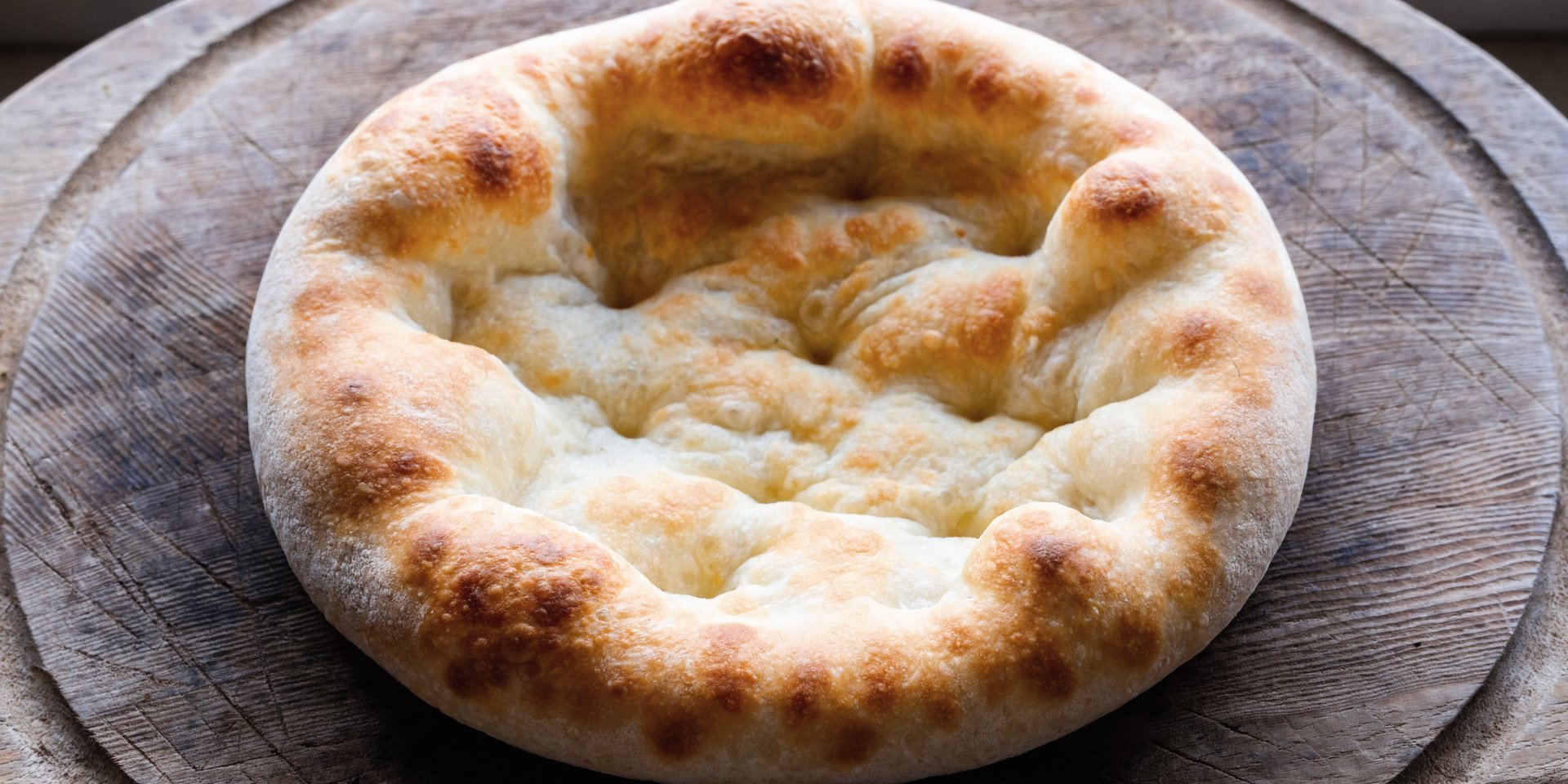 A close-up, top-down view of a golden-brown pizza crust with airy edges on a wooden surface.