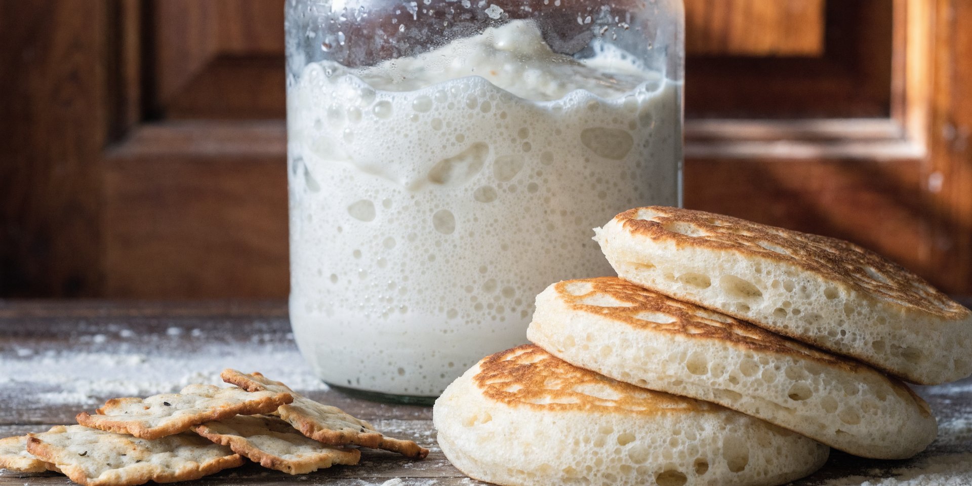 A close-up photograph shows a glass jar of sourdough starter in the background and a pile of sourdough discard crackers and pancakes in the foreground.