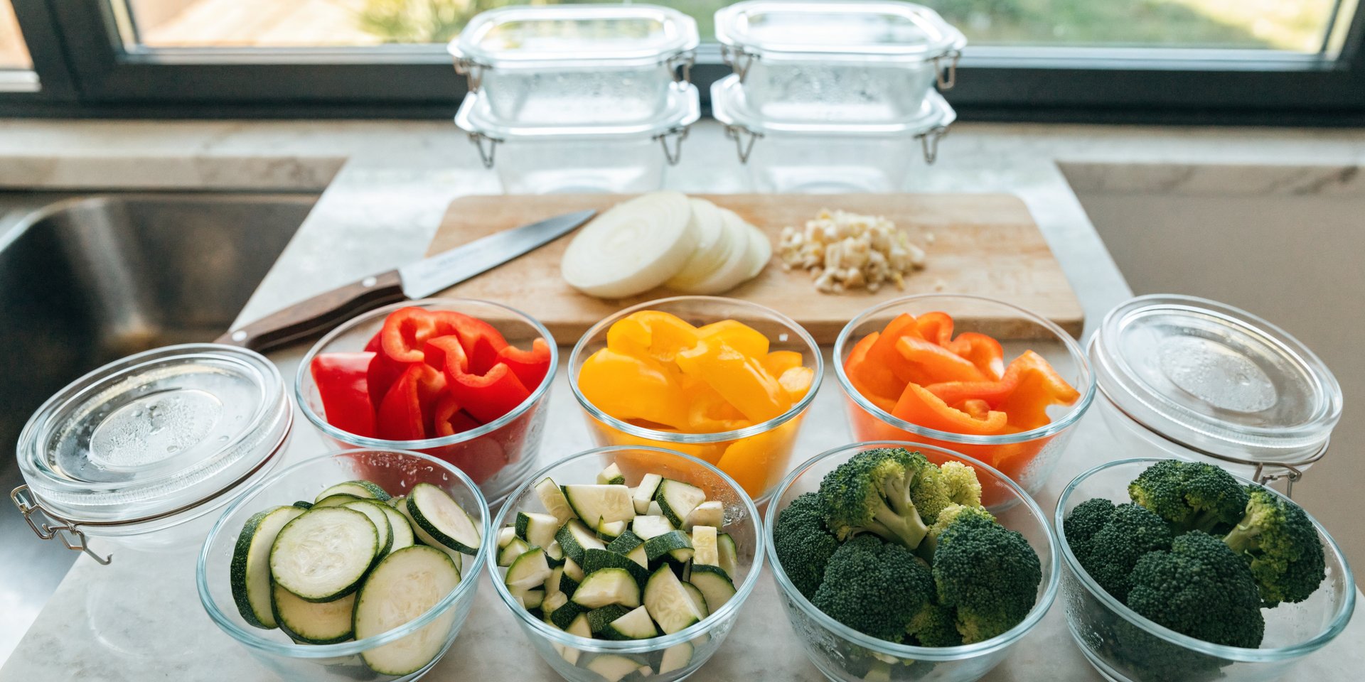 An overhead shot of a kitchen counter with various prepped vegetables, including chopped bell peppers, zucchini, broccoli, onion, and garlic, in glass containers and on a cutting board.