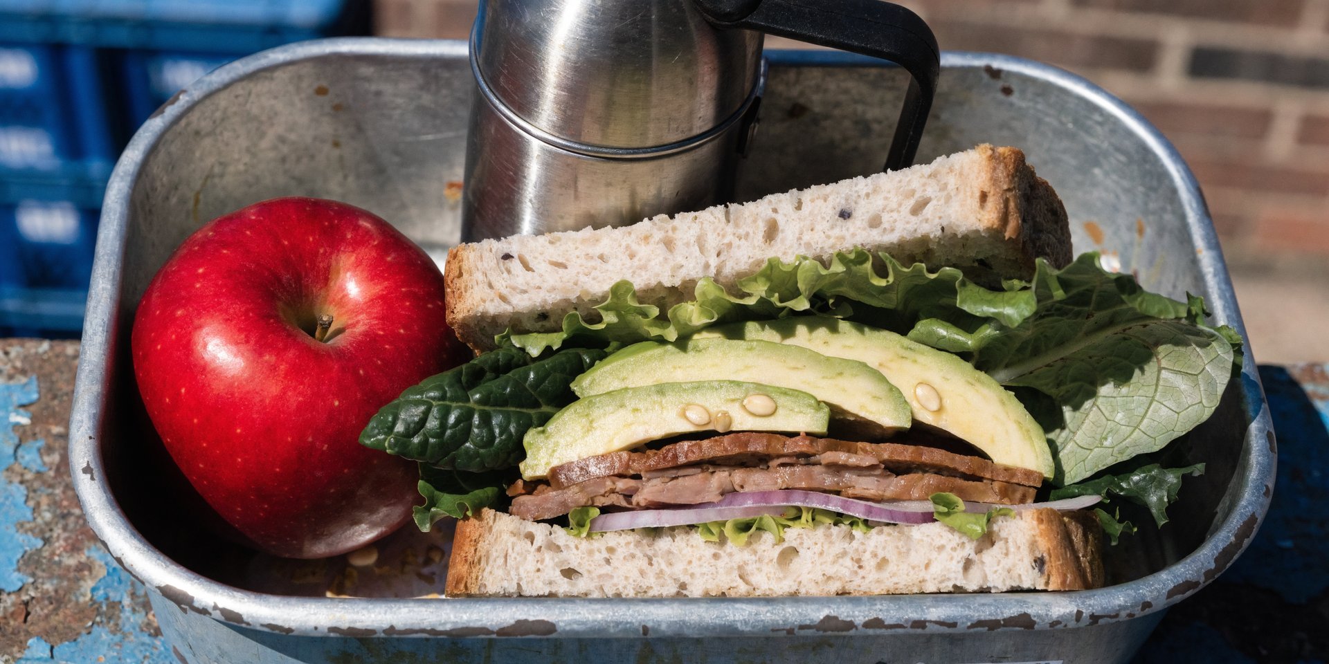 A thick turkey and avocado sandwich in a lunchbox with an apple and a thermos.