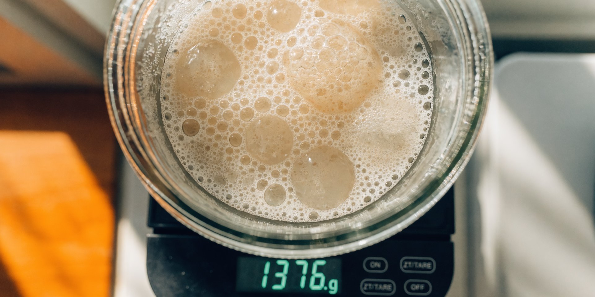 A digital kitchen scale weighing a glass jar filled with bubbly sourdough discard, displaying a gram measurement.