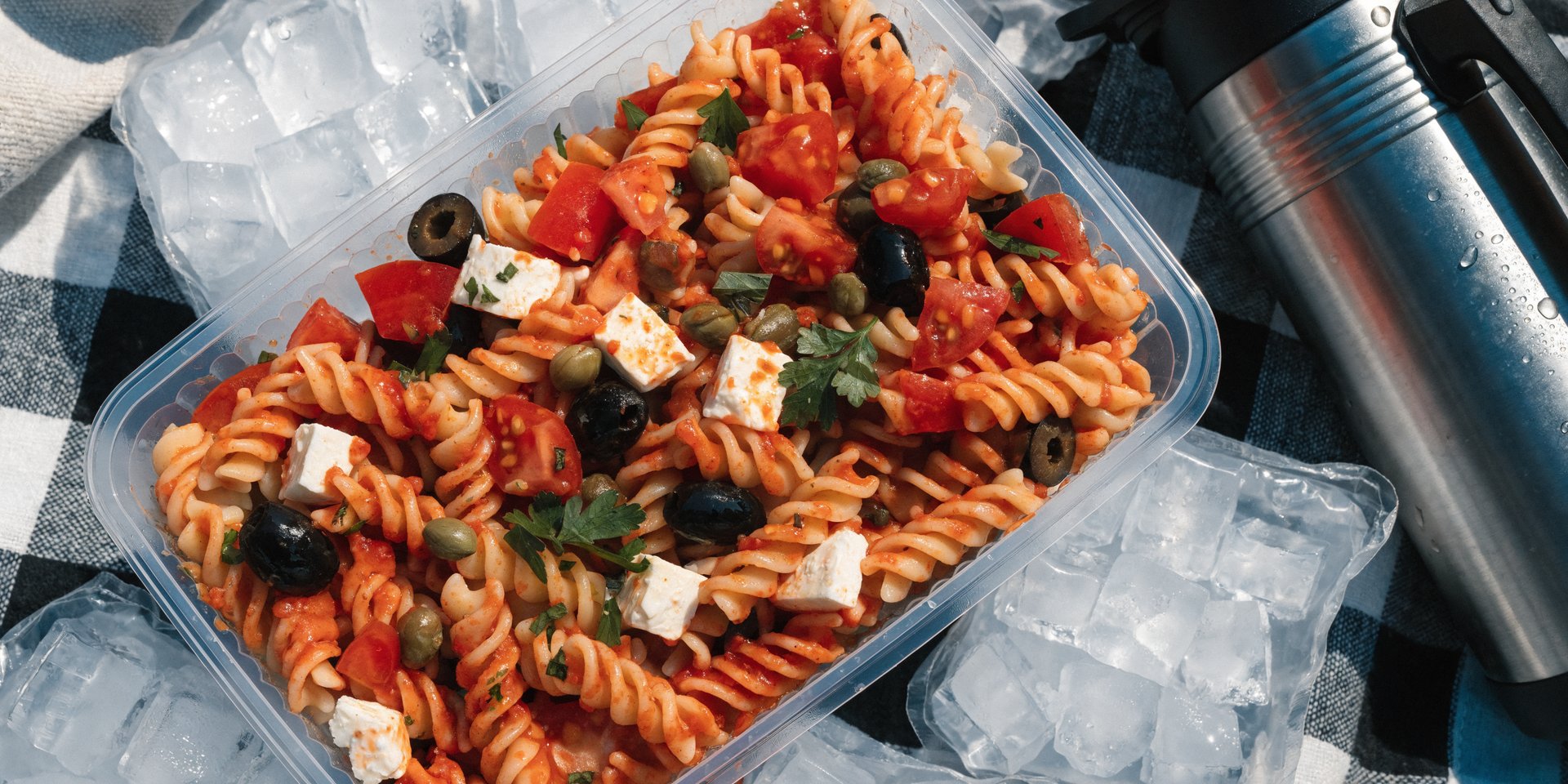 A close-up overhead shot of a colorful rotini pasta salad packed in a clear container for a picnic.