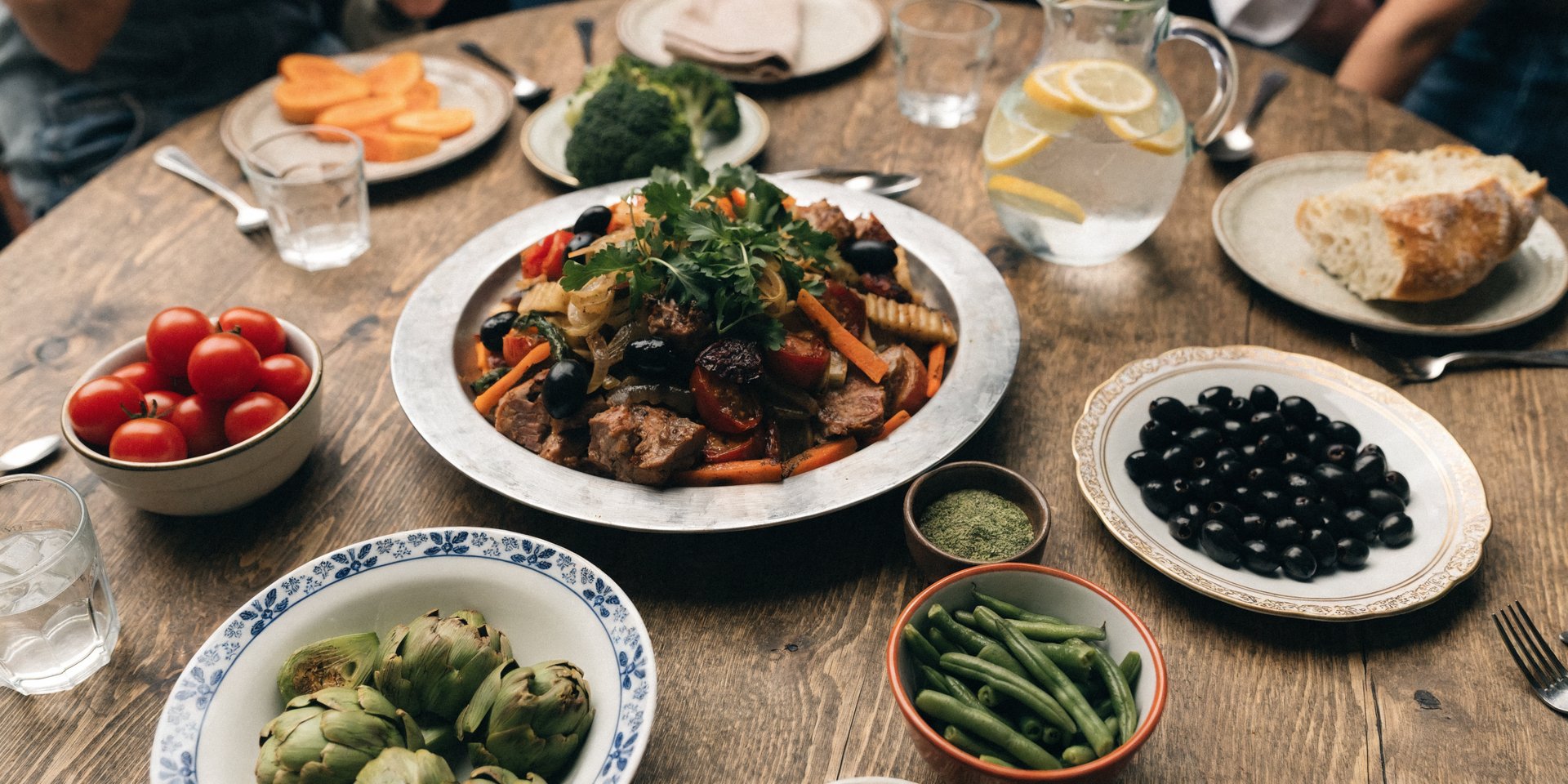 An overhead shot of a large, rustic wooden table filled with a colorful, abundant family-style meal, including a central casserole dish and various side dishes.