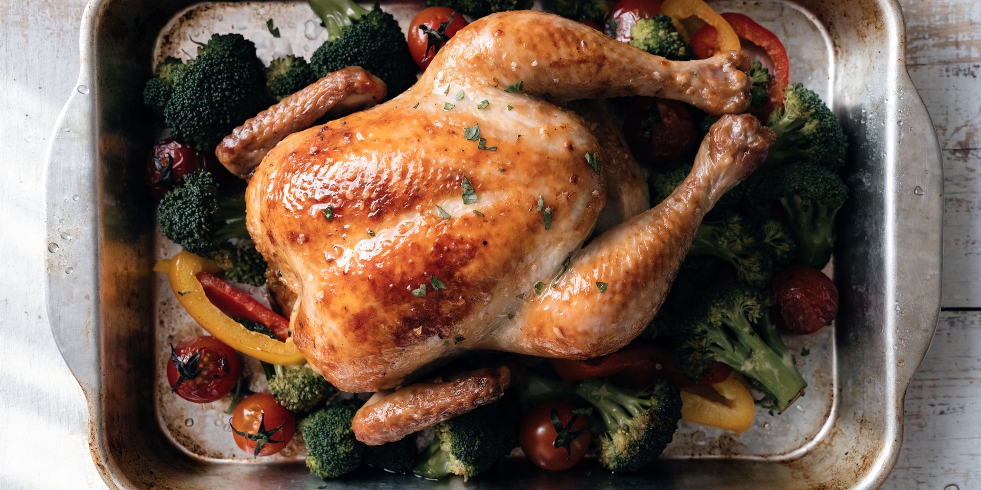A brightly lit overhead shot of a sheet pan filled with roasted chicken and various vegetables, including broccoli, bell peppers, and cherry tomatoes, garnished with fresh herbs.