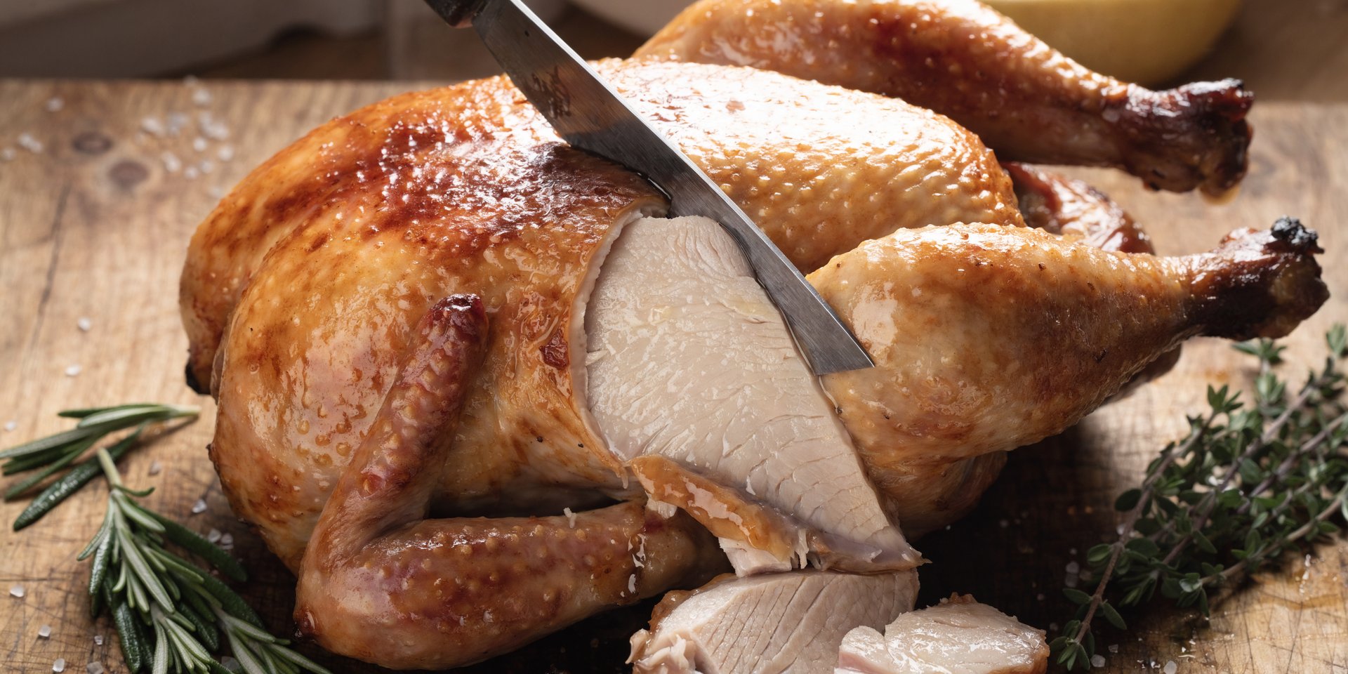 A close up of a carved rotisserie chicken on a wooden cutting board with a knife and herbs.
