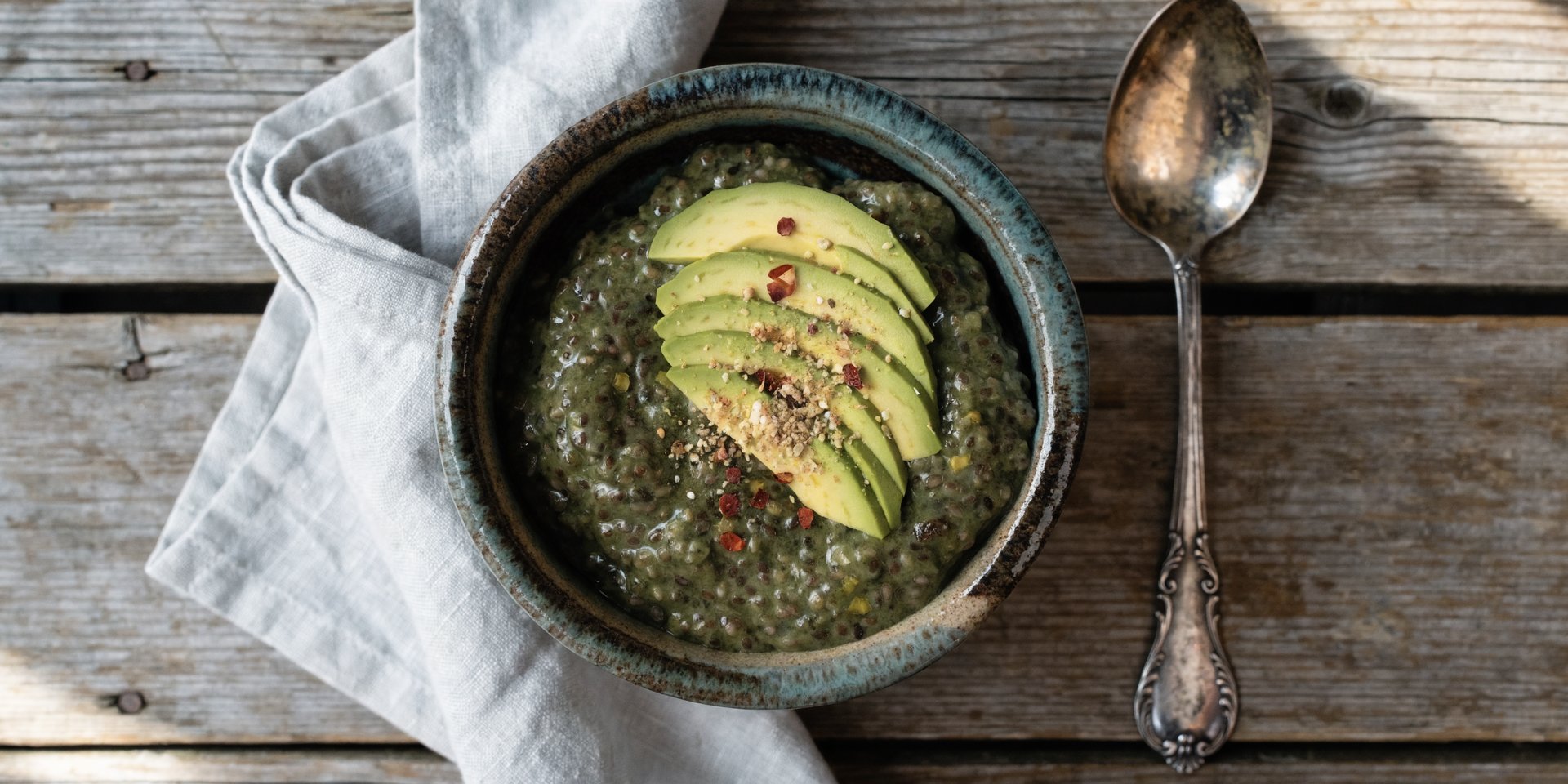 An overhead shot of a green savory chia pudding in a rustic bowl, garnished with avocado slices, everything bagel seasoning, and chili flakes.