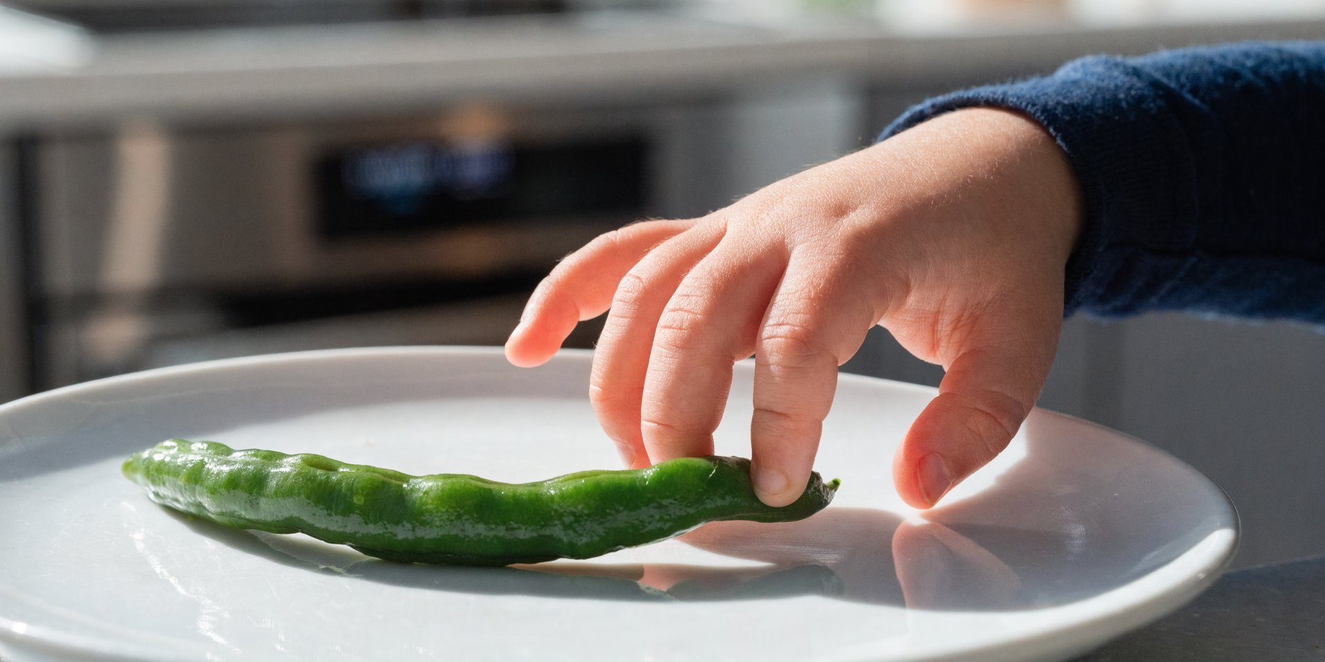 A child's hand reaches for a single green bean on a white plate.